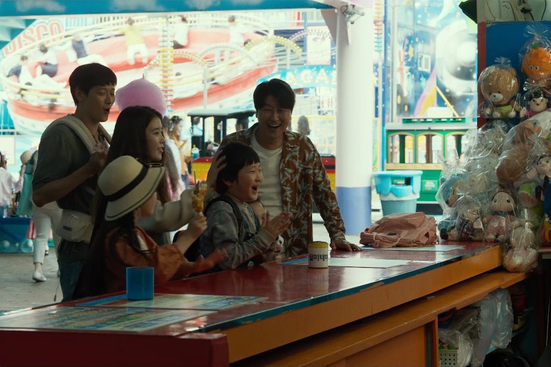 Grupa osób, w tym dwójka dzieci i troje dorosłych, stoi podekscytowana przy kolorowym stoisku z grami karnawałowymi, gdzie nagrodami są pluszowe zabawki, uśmiecha się i wiwatuje w ożywionej atmosferze parku rozrywki.