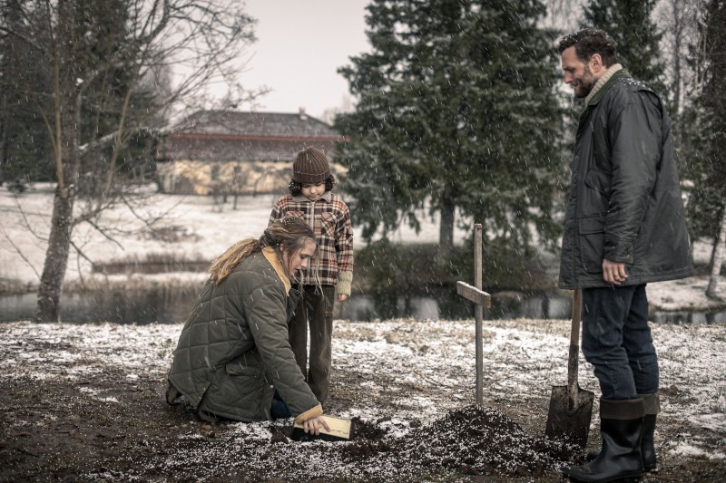 Kobieta klęczy przy małym grobie oznaczonym drewnianym krzyżem, kładąc przedmiot na ziemi, podczas gdy dziecko i mężczyzna z łopatą stoją w pobliżu. Pada śnieg, a w tle widać drzewa.