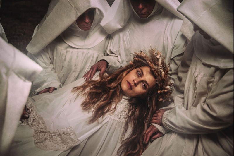 Kobieta z długimi włosami w białej sukni i koronie z suszonych kwiatów leży w ramionach ludzi ubranych w białe szaty i kaptury, ich twarze są w większości ukryte. Scena jest ponura i eteryczna.