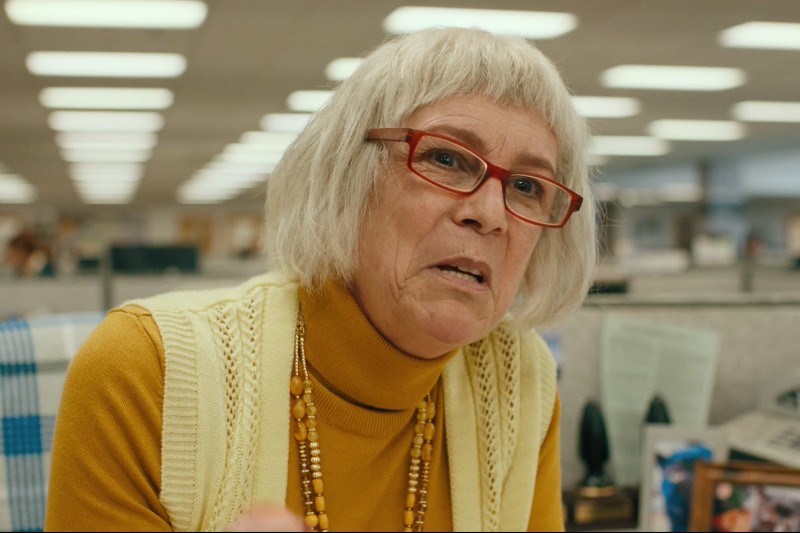 Starsza kobieta z krótkimi siwymi włosami i czerwonymi okularami, ubrana w żółty golf i kremową kamizelkę, siedzi w biurze z jarzeniówkami i boksami w tle.