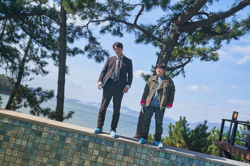 Dwie osoby stoją na skraju kafelkowej konstrukcji w pobliżu zbiornika wodnego, nosząc ochronne niebieskie ochraniacze na buty. Jedna jest w garniturze, druga w swobodnym ubraniu outdoorowym. W tle widać sosny i odległy ląd.