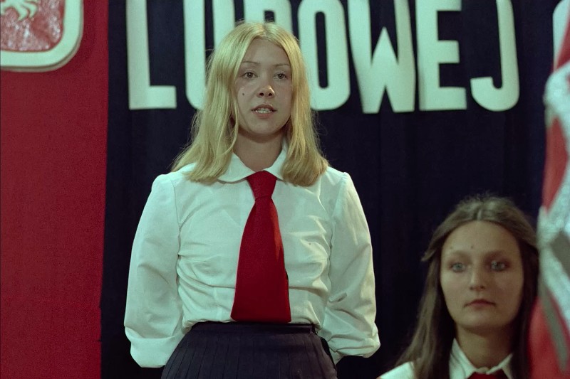 Młoda kobieta o blond włosach, ubrana w białą koszulę, czerwony krawat i ciemną spódnicę stoi przed czerwono-czarnym tłem z dużymi białymi literami. Inna kobieta o długich brązowych włosach siedzi obok niej.