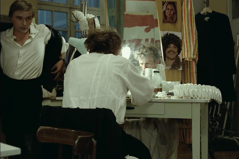 Dwie osoby są w garderobie. Jedna siedzi przy zagraconym stole, patrzy w lustro i szykuje się, podczas gdy druga poprawia marynarkę. W tle widać zdjęcia i kostiumy.