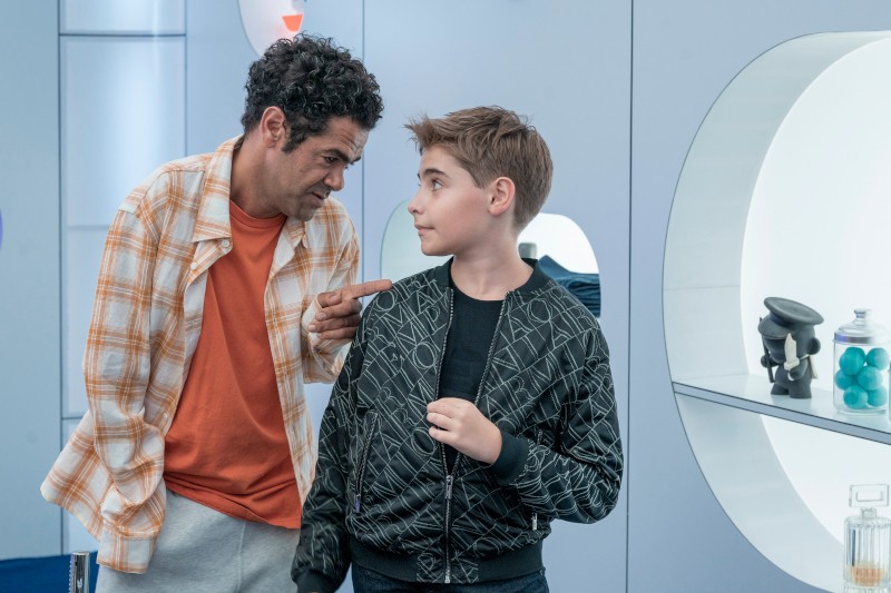 Dorosły mężczyzna w pomarańczowej koszuli gestykuluje i rozmawia z młodym chłopcem w wzorzystej czarnej kurtce. Znajdują się w nowoczesnym, futurystycznym pokoju z półkami i dekoracyjnymi przedmiotami.