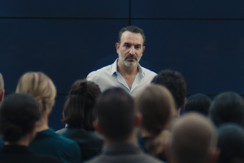 Mężczyzna o poważnym wyrazie twarzy stoi przed grupą siedzących ludzi, sprawiając wrażenie, że wygłasza przemówienie lub prezentację. Ma na sobie jasną koszulę i stoi twarzą do publiczności, którą widać od tyłu.