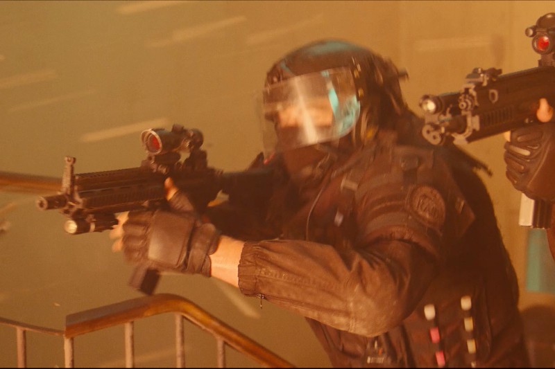 Osoba w stroju taktycznym i z przezroczystą osłoną twarzy trzyma karabin, wspinając się po schodach z inną uzbrojoną osobą za sobą. Scena jest napięta, z dramatycznym oświetleniem sugerującym akcję lub konflikt.