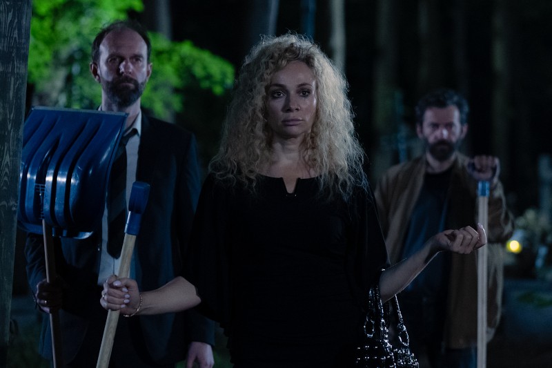 Trzy osoby stoją na zewnątrz nocą, dwóch mężczyzn trzyma łopaty, a kobieta z kręconymi blond włosami w środku, ubrana na czarno i niosąca torebkę, otoczona drzewami. Atmosfera jest napięta i tajemnicza.