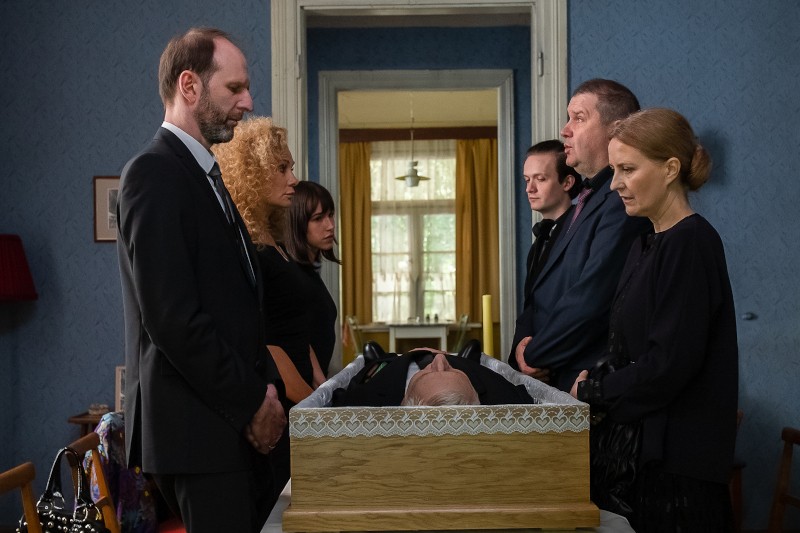 Sześć osób ubranych w ciemne, formalne stroje stoi wokół drewnianej trumny w pomieszczeniu, opłakując osobę w środku. Atmosfera jest ponura, a w pokoju są niebieskie tapety i oprawione zdjęcia na ścianach.