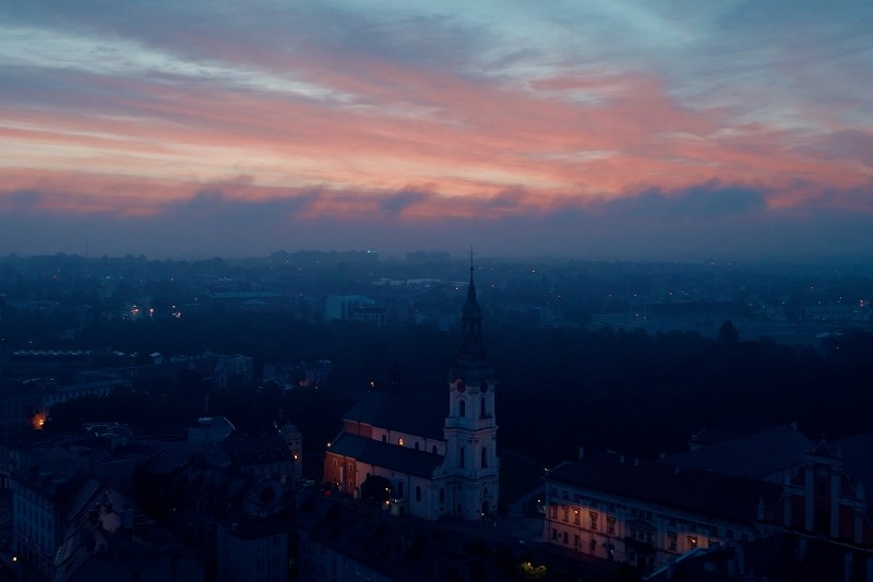 Krajobraz miejski o zmierzchu z kościołem z wysoką iglicą na pierwszym planie, otoczonym budynkami. Niebo wypełniają niebieskie i różowe chmury, gdy zachodzi słońce, rzucając miękkie światło na scenę.
