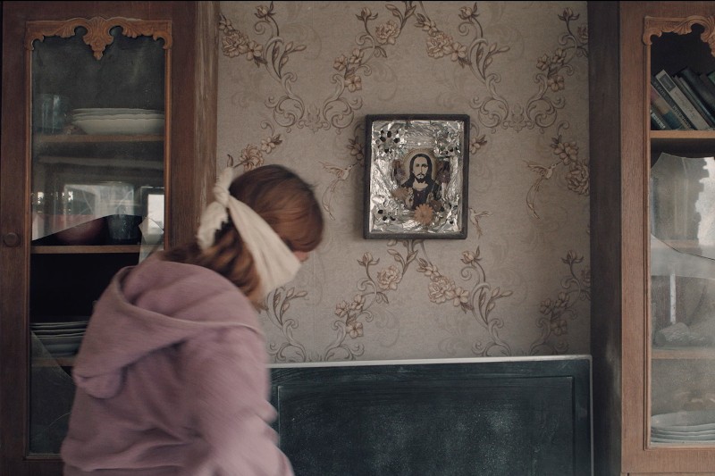 Osoba o jasnobrązowych włosach, z zawiązanymi oczami i w fioletowym kapturze, stoi przed ścianą z kwiatową tapetą i oprawioną ikoną religijną, otoczoną dwiema przeszklonymi gablotami.
