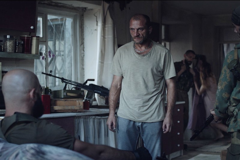 Napięta scena w słabo oświetlonym, zagraconym pokoju pokazuje mężczyznę w beżowej koszuli stojącego i wpatrzonego w niego uważnie, w pobliżu uzbrojonych mężczyzn i parę obejmującą się w tle. Broń jest widoczna na stole i w rękach ludzi.