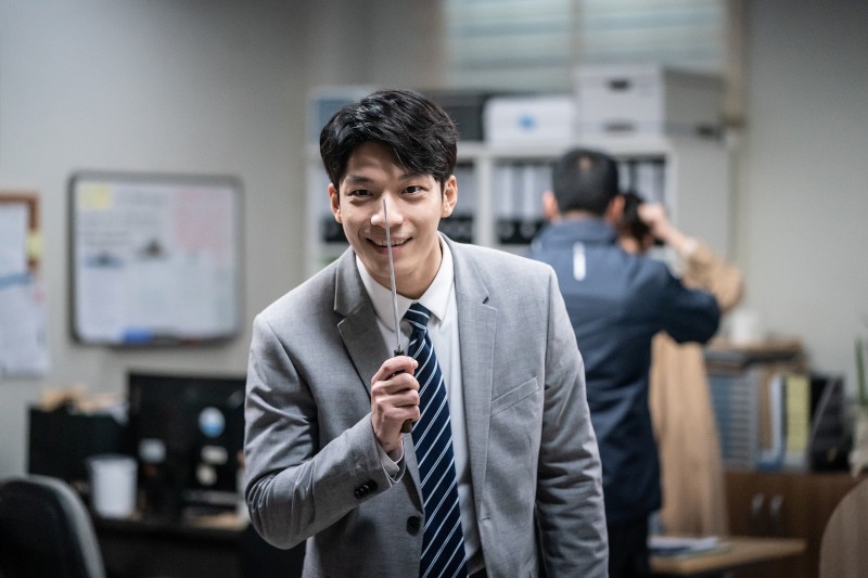 Młody mężczyzna w szarym garniturze i pasiastym krawacie uśmiecha się, trzymając żartobliwie długopis przed twarzą w biurze. Dwie osoby są rozmazane w tle przy półkach i tablicy ogłoszeń.