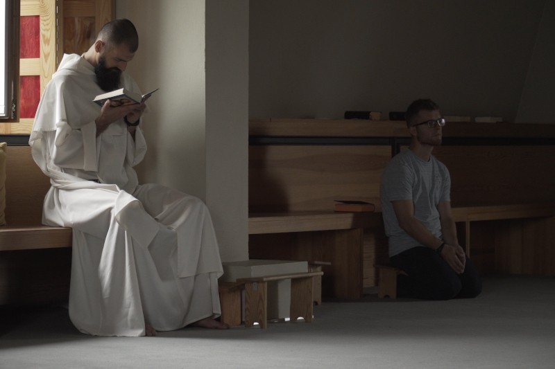 Mężczyzna ubrany w białą szatę czyta książkę, siedząc na drewnianej ławce; inny mężczyzna ubrany w zwykłe ubranie klęczy obok i modli się w słabo oświetlonym pomieszczeniu.