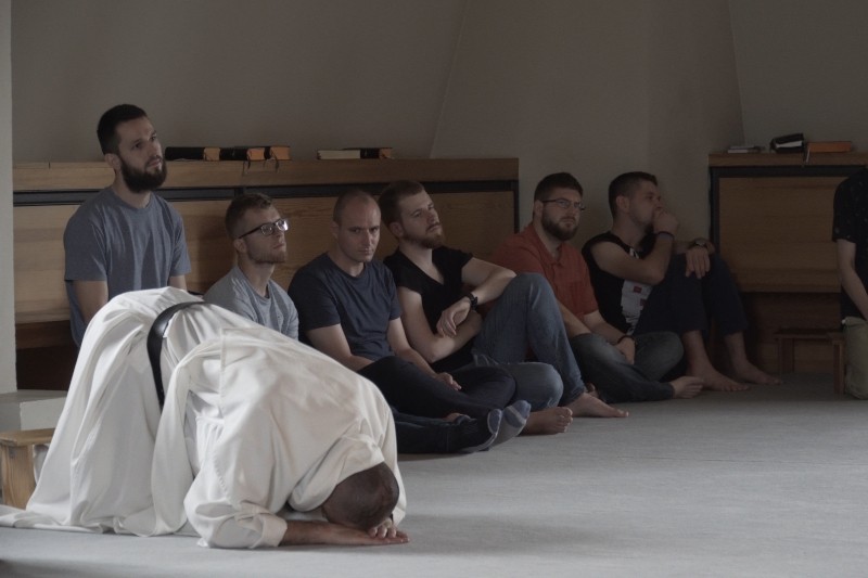 Grupa mężczyzn siedzi boso na ławkach i podłodze, obserwując osobę w białej szacie, która pochyla się nisko, dotykając czołem ziemi w cichym, zamkniętym pomieszczeniu.