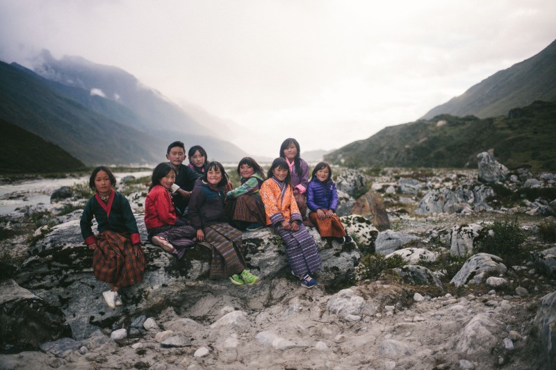 Grupa dzieci ubranych w kolorowe ubrania siedzi na skałach na tle górzystego krajobrazu, w tle widać rzekę i mgliste wzgórza.