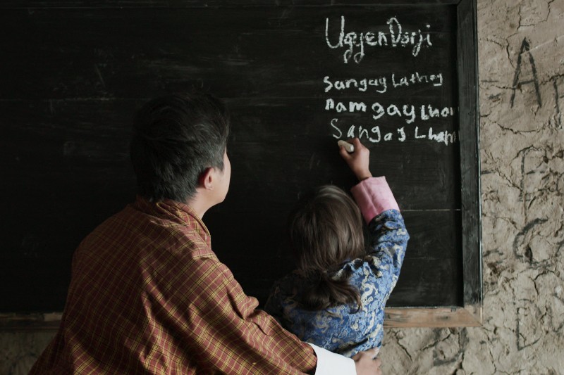 Nauczyciel pomaga młodemu uczniowi pisać słowa kredą na tablicy w rustykalnej klasie z teksturowanymi ścianami. Oboje noszą tradycyjne stroje i stoją twarzą do tablicy.