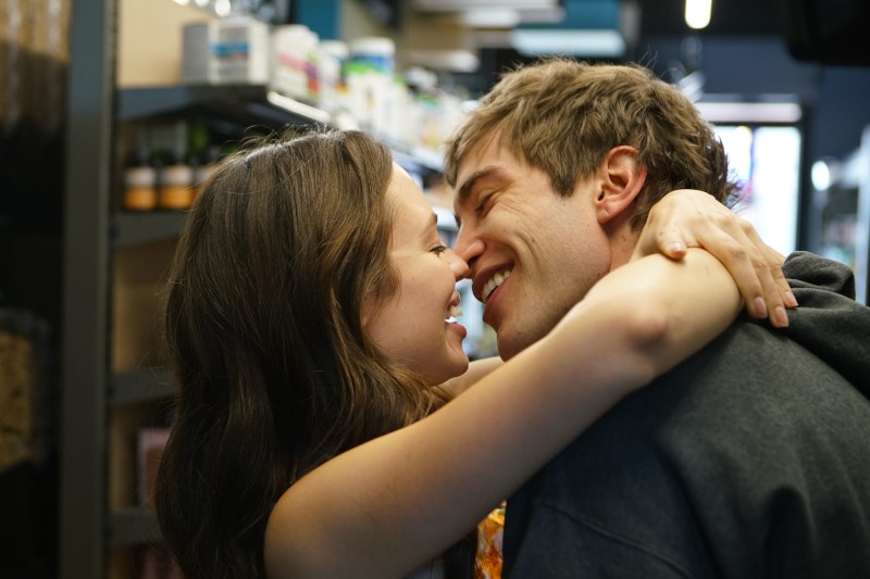 Młoda para stoi blisko siebie w alejce sklepowej, uśmiechając się i obejmując czule, ich twarze prawie się stykają, wyglądając na szczęśliwych i pełnych uczuć. W tle widoczne są półki z produktami.