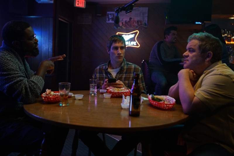 Trzech mężczyzn siedzi przy okrągłym stole w słabo oświetlonym barze, przed nimi stoją drinki i kosze z jedzeniem. Jeden z nich gestykuluje, mówiąc, drugi patrzy na telefon, a trzeci słucha, opierając rękę na brodzie.
