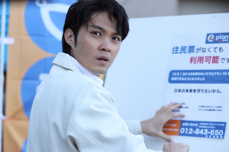 Mężczyzna w białej kurtce wygląda na zaskoczonego, dotykając znaku z japońskim tekstem i numerem telefonu. Tło obejmuje niebieski i pomarańczowy baner.