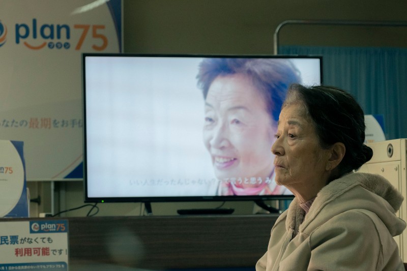 Starsza kobieta siedzi w poczekalni, wyglądając na zamyśloną. Za nią ekran telewizora wyświetla inną uśmiechniętą starszą kobietę z nałożonym japońskim tekstem. W pomieszczeniu widoczne są znaki Planu 75.