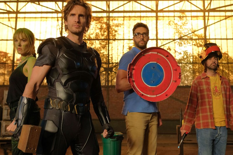 Cztery osoby w różnych kostiumach stoją w oświetlonym słońcem magazynie: jedna w czarnej zbroi, druga trzyma czerwoną tarczę, trzecia w zielonej farbie na ciele i trzecia w flanelowej koszuli z hełmem i prowizoryczną bronią.