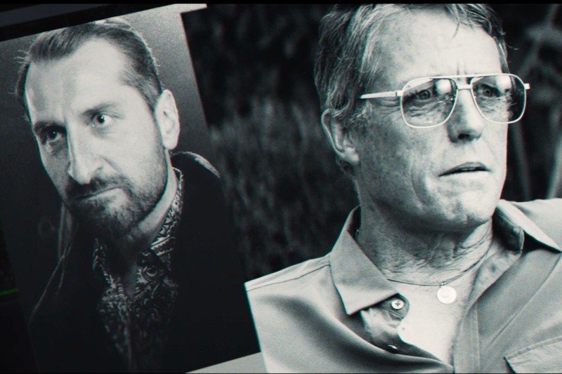 Czarno-biały obraz przedstawiający mężczyznę w okularach przeciwsłonecznych i koszuli z kołnierzykiem, siedzącego obok wydrukowanego zdjęcia innego mężczyzny z brodą i intensywnym wyrazem twarzy.