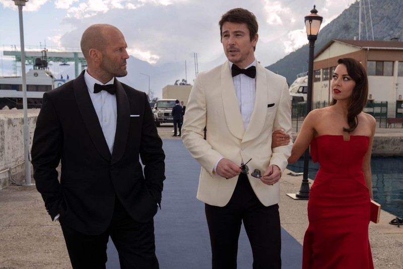 Trzy osoby w strojach formalnych idą razem na zewnątrz, wzdłuż brzegu wody; mężczyzna po lewej stronie ma na sobie czarny smoking, mężczyzna w środku białą marynarkę smokingową, a kobieta po prawej ma na sobie czerwoną suknię bez ramiączek.