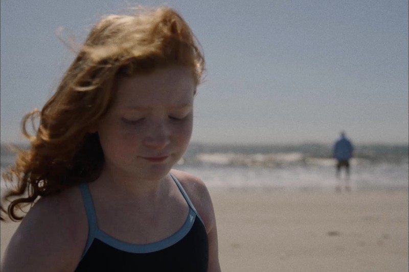 Młoda dziewczyna z rudymi włosami w kostiumie kąpielowym stoi na słonecznej plaży, patrząc w dół z zamyślonym wyrazem twarzy. Ocean i odległa postać są rozmazane w tle.