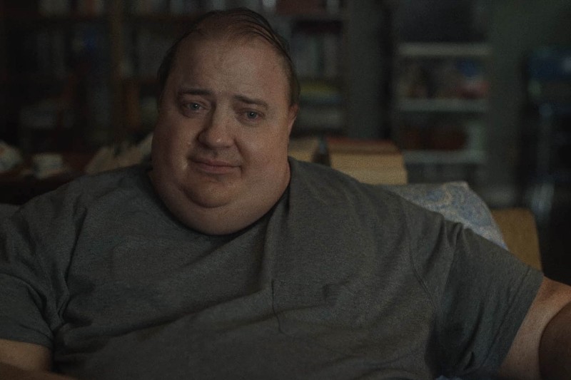 Mężczyzna z krótkimi brązowymi włosami i szarym T-shirtem siedzi w środku, patrząc prosto przed siebie z łagodnym, emocjonalnym wyrazem twarzy. Na delikatnie oświetlonym tle widać regał z książkami i stos książek.