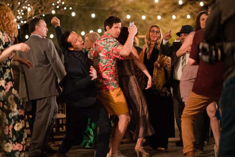 Ludzie energicznie tańczą na imprezie plenerowej pod lampkami. Jeden mężczyzna w koszuli w kwiaty i krótkich spodenkach wyróżnia się w środku. Tłum wydaje się radosny i odświętny, ubrany w mieszankę strojów swobodnych i formalnych.