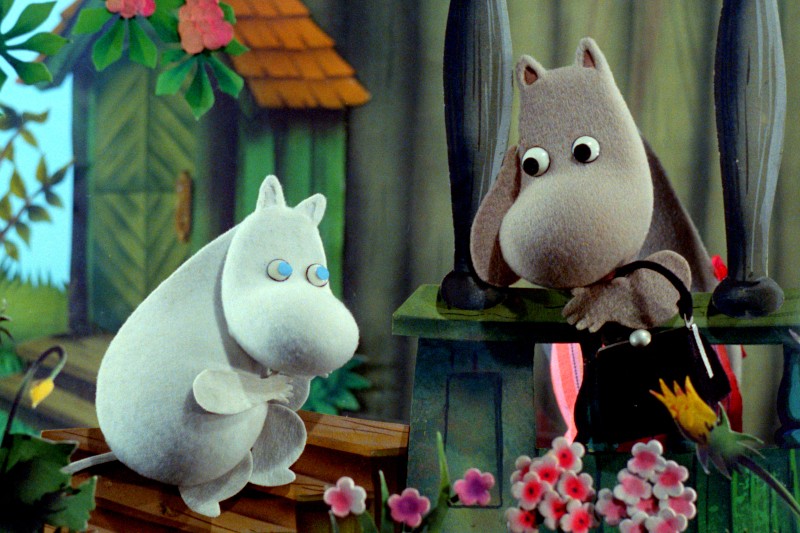 Dwie pluszowe postacie, jedna biała i jedna szara, znajdują się w kolorowym ogrodzie z kwiatami. Szara postać zagląda do czarnej torebki, podczas gdy biała postać siedzi na drewnianych schodach, obie z zaskoczonymi minami.