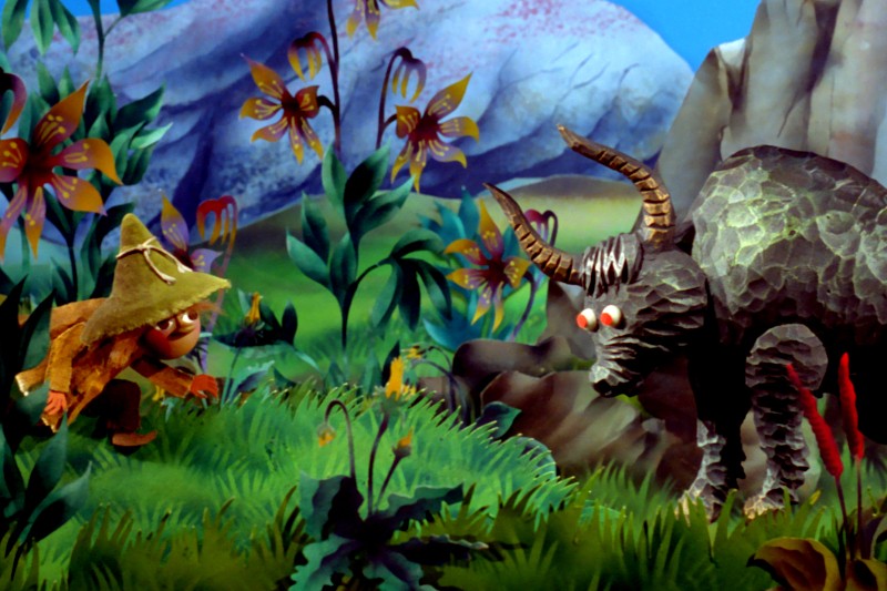 Mała postać w słomkowym kapeluszu zbliża się do dużego, rogatego stworzenia o czerwonych oczach na tle barwnego, bujnego i kapryśnego krajobrazu z kwiatami, zielenią i górami w tle.