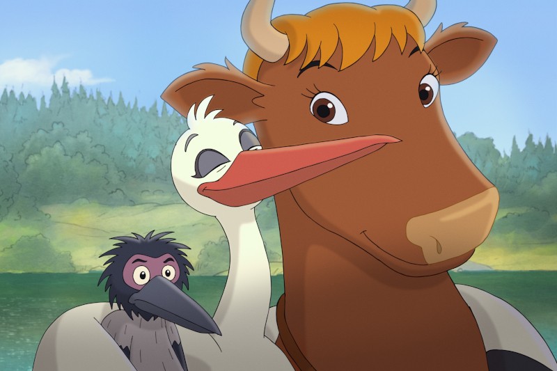 Przyjazna, animowana, brązowa krowa uśmiecha się obok białego bociana i małego czarno-szarego ptaka, na tle drzew i błękitnego nieba.