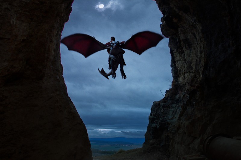 Osoba jadąca na dużym, czarnym smoku o czerwonych skrzydłach wylatuje nocą z jaskini, na tle zachmurzonego nieba i pełni księżyca.