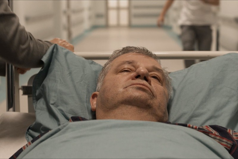 Starszy mężczyzna leży na szpitalnym łóżku, patrzy w górę ze zmęczonym wyrazem twarzy. Ktoś, kogo ręka jest częściowo widoczna, wiezie go na szpitalnym korytarzu.