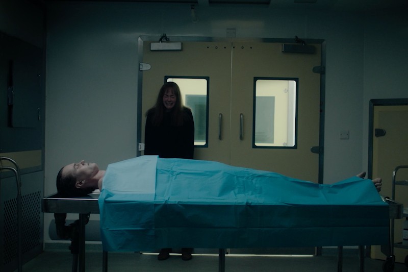 Osoba przykryta niebieskim prześcieradłem leży na stole w kostnicy, a kobieta stoi za ciałem, wyglądając na przerażoną, w słabo oświetlonym pomieszczeniu z podwójnymi drzwiami w tle.