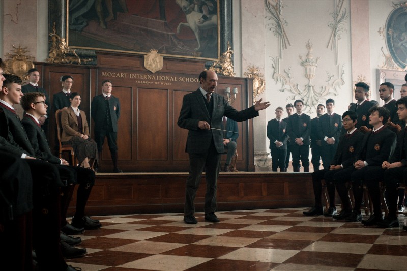 Nauczyciel stoi i gestykuluje, zwracając się do uczniów w mundurkach, siedzących w wielkiej sali z szachownicą na podłodze w Mozart International School. Inni uczniowie i kadra pedagogiczna obserwują ich z tła.