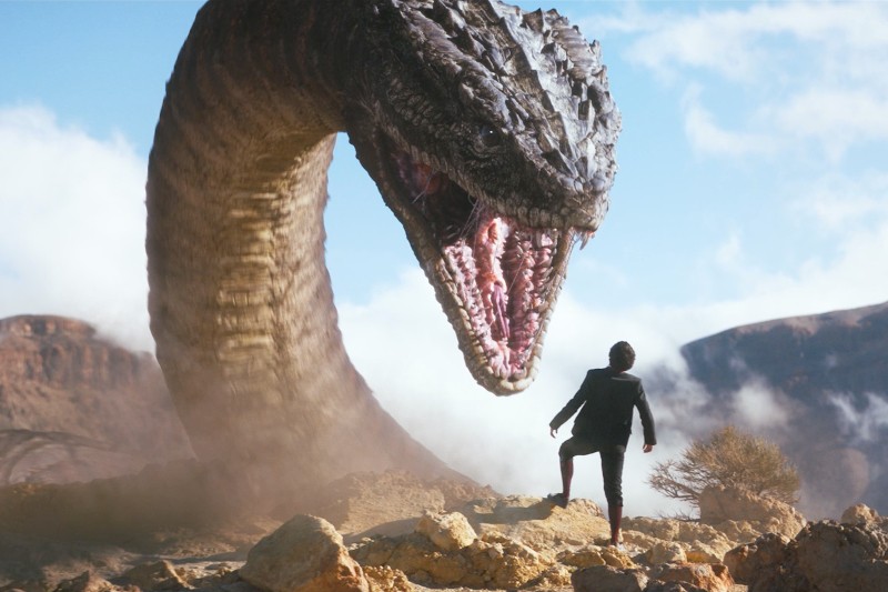 Osoba w garniturze stoi na skalistym terenie naprzeciwko ogromnego, górującego nad resztą węża z szeroko otwartą paszczą, odsłaniającą ostre zęby. Obraz na tle malowniczego nieba i pustynnego krajobrazu.