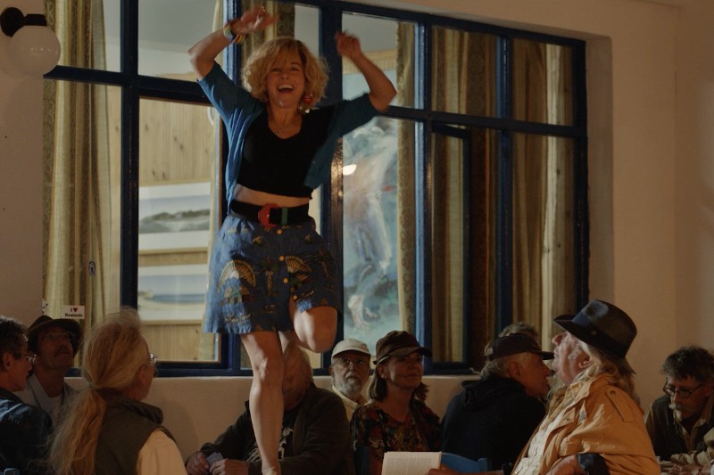 Kobieta w niebieskiej spódnicy i kardiganie energicznie tańczy na stole w zatłoczonym pomieszczeniu, podczas gdy ludzie siedzący wokół niej patrzą i się uśmiechają. Sceneria wydaje się świąteczna i pełna życia.