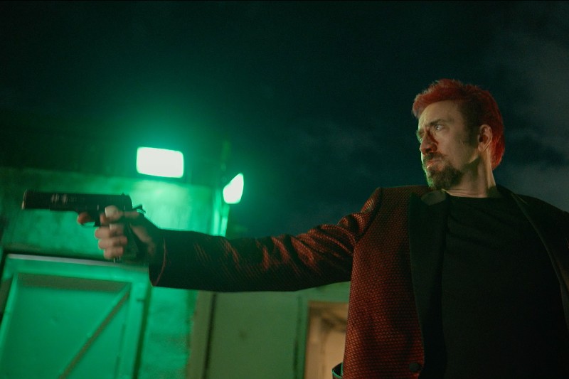Mężczyzna z rudymi włosami i brodą, ubrany w ciemnoczerwoną kurtkę, w nocy celuje pistoletem w lewo. Stoi przed budynkiem oświetlonym zielonym światłem. Scena wydaje się napięta i dramatyczna.
