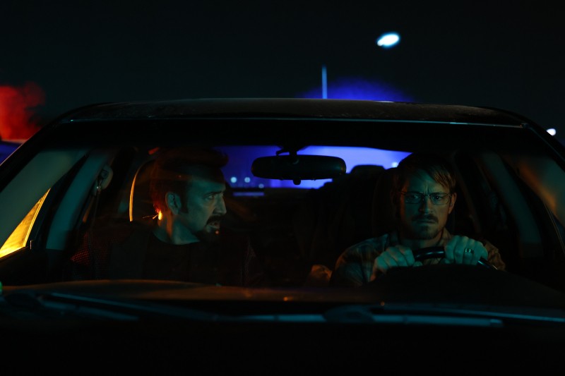 Dwóch mężczyzn siedzi w samochodzie nocą; kierowca wygląda na napiętego i trzyma kierownicę, podczas gdy pasażer patrzy na niego. W tle migają policyjne światła, rzucając niebieskie i czerwone światło na samochód.