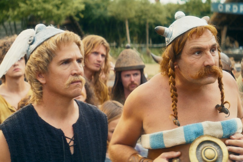 Dwóch mężczyzn przebranych za postacie komiksowe Asterix i Obelix, ubranych w hełmy i kostiumy, stoi na zewnątrz wśród tłumu, patrząc przed siebie z poważnymi minami. W tle widać drzewa i rozmazanych ludzi.