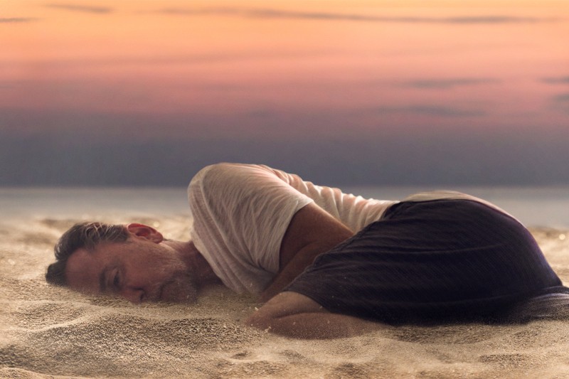 Mężczyzna w białej koszulce i ciemnych spodenkach leży zwinięty na boku na piaszczystym gruncie na plaży podczas zachodu słońca, w tle widać spokojne morze i kolorowe niebo.