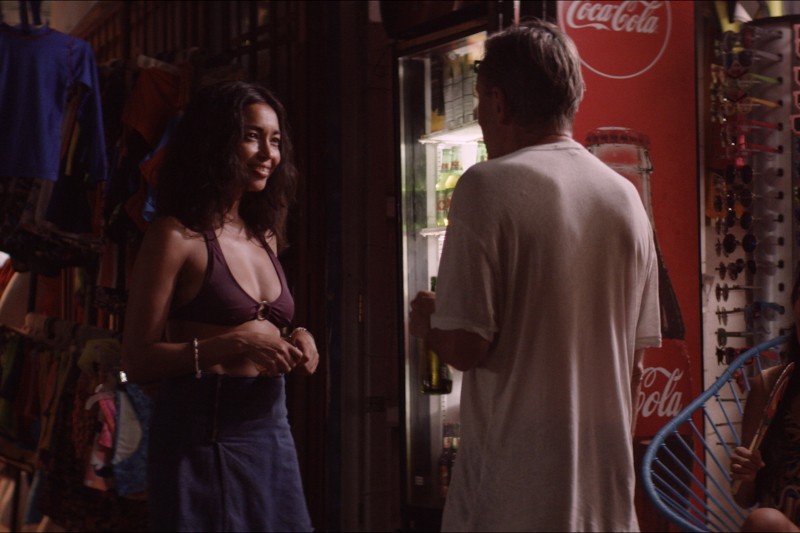 Kobieta w fioletowym staniku od bikini i niebieskiej spódnicy rozmawia z mężczyzną w białym T-shircie, stojącym obok lodówki pełnej napoi, w tle widać czerwony znak Coca-Coli i różne przedmioty.