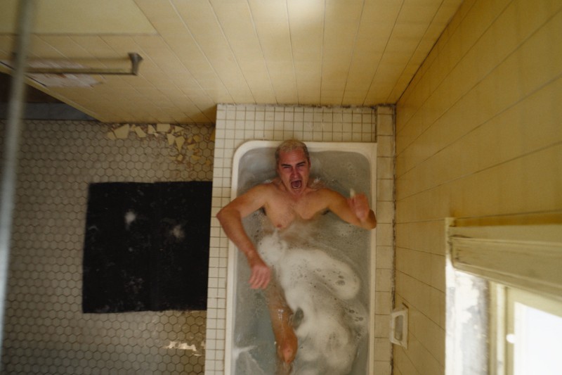 Mężczyzna o blond włosach leży w małej wannie wypełnionej mętną wodą, patrząc w górę z zaniepokojonym wyrazem twarzy. Łazienka wygląda na starą, z pożółkłymi kafelkami i łuszczącą się farbą na ścianie.