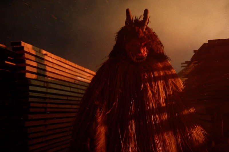 Osoba ubrana w duży, pokryty słomą kostium z rogatą, czerwoną maską na twarzy stoi groźnie wśród drewnianych desek w dramatycznym, zadymionym oświetleniu.