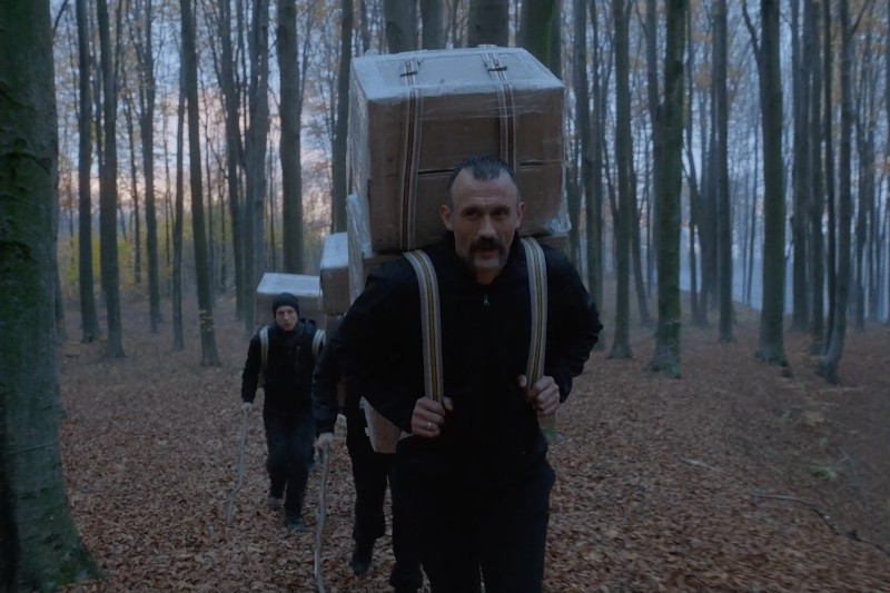 Trzy osoby idą przez las, każda niosąca duży, pudełkowaty pakunek przytroczony do pleców. Ziemia jest pokryta opadłymi liśćmi, a drzewa są wysokie i nagie, co sugeruje późną jesień lub wczesną zimę.
