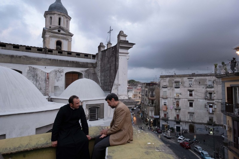 Dwóch mężczyzn, jeden w szacie kapłańskiej, a drugi w płaszczu, siedzi i rozmawia na dachu, z którego roztacza się widok na stare europejskie miasto ze zwieńczonymi kopułami budynkami i zachmurzonym niebem w tle.