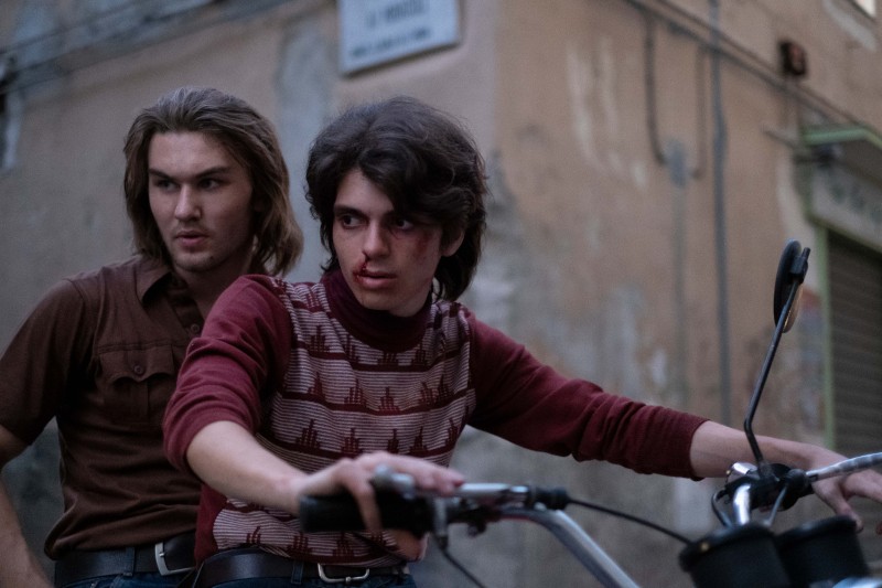 Dwóch młodych mężczyzn z długimi włosami jedzie razem na rowerze przez zaniedbaną ulicę miasta; mężczyzna z przodu ma posiniaczoną, zakrwawioną twarz i wygląda na napiętego, podczas gdy mężczyzna jadący za nim wygląda na zaniepokojonego.
