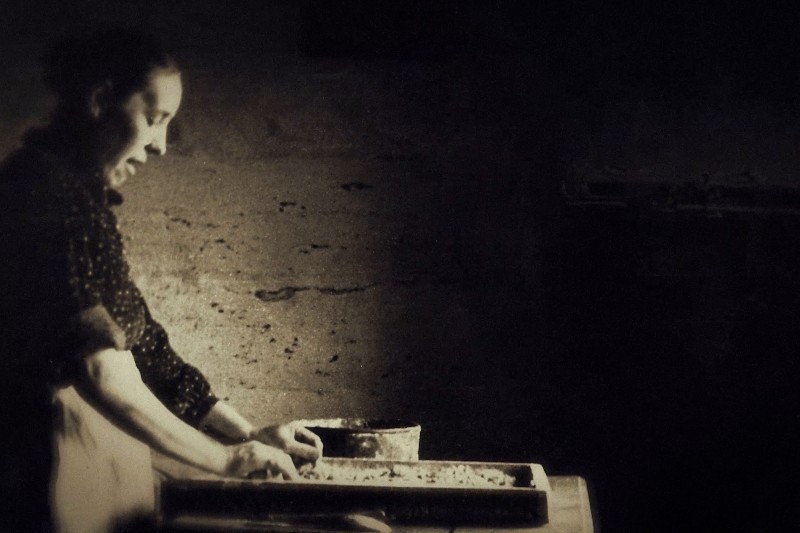 Kobieta w ciemnym, zabytkowym otoczeniu stoi przy blacie roboczym, formując ciasto lub wypieki na tacy rękoma. Nosi ciemną sukienkę z podwiniętymi rękawami i fartuch, a częściowo oświetla ją miękkie światło.